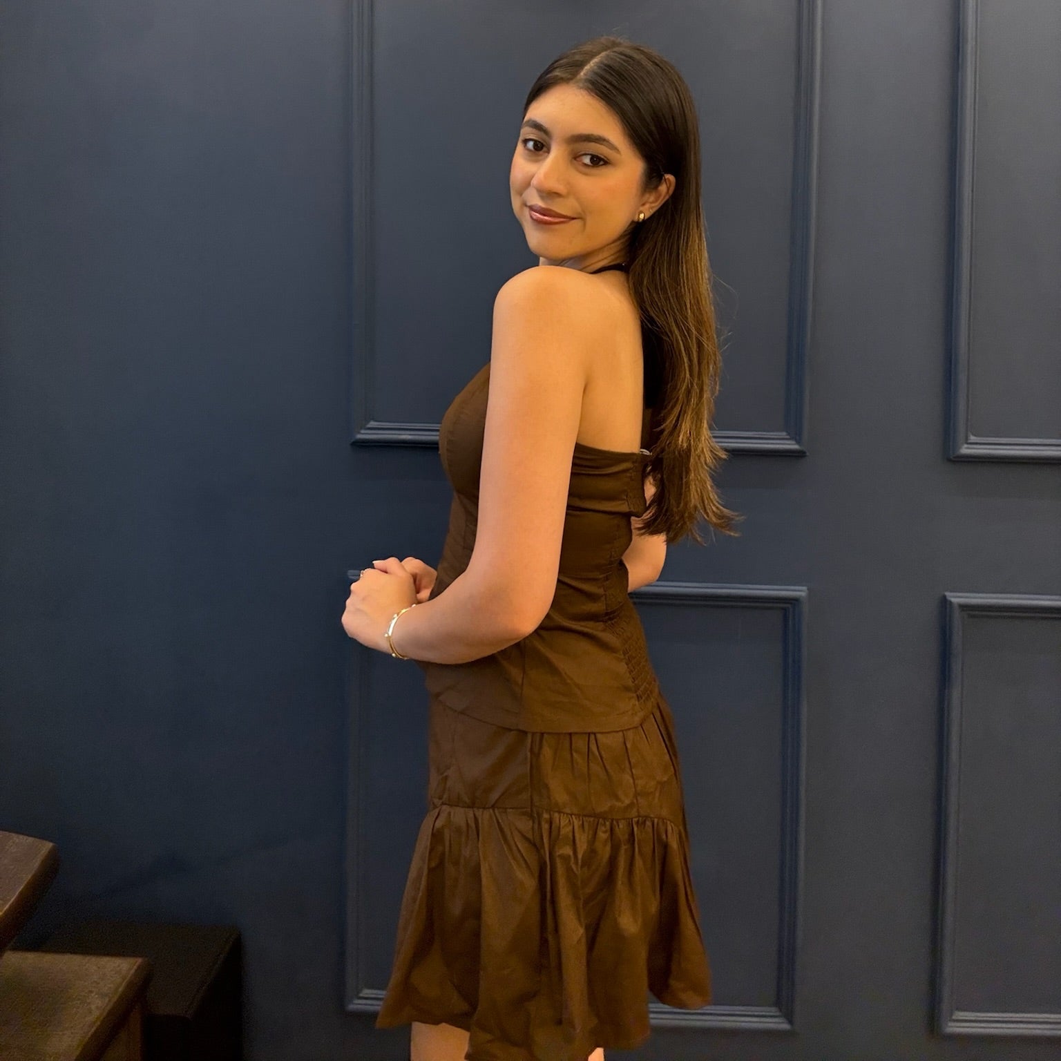 Woman in a brown dress standing against a dark blue paneled wall.